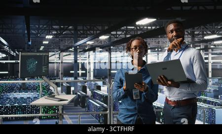 Data center admins running diagnostics on equipment using system monitoring tools to identify errors, doing brainstorming. IT experts work together to prevent failure and minimize downtime, Stock Photo