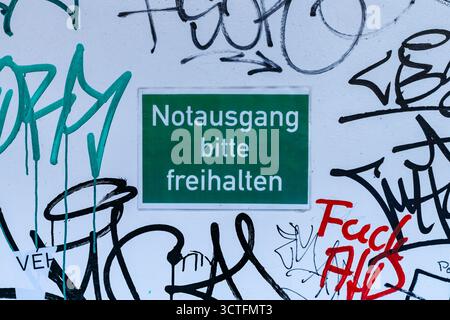 Green emergency exit sign (Notausgang) with graffiti on a white wall, close-up urban detail, Berlin, Germany Stock Photo