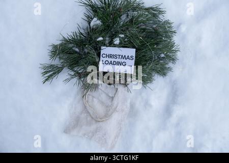 Paper note with text CHRISTMAS LOADING on Reusable shopping bag with fir spruce green branches in snowy outdoor winter forest background. Merry Christmas and new year concept. Natural bouquet greeting card copy space. Garland lights Stock Photo