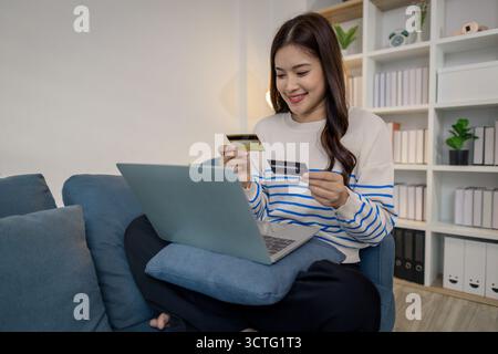A young woman sits on a sofa using a laptop while holding credit cards, representing secure online payment and modern lifestyle. She smiles confidentl Stock Photo