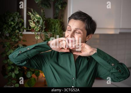 Satisfied mature woman touching soft skin and stretching in morning on home kitchen, self care love Stock Photo