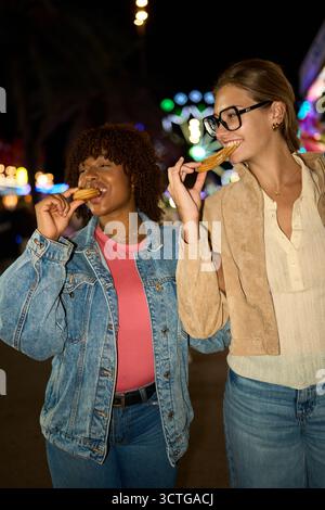 Two women friends enjoying churros at night fairground Stock Photo