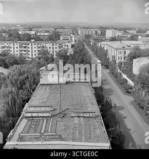 A high-angle rooftop view looking down on Svobody Street in Sloviansk, Ukrainian SSR, during the 1970s. The panoramic photograph captures a peaceful cityscape from the Brezhnev era of stagnation. The street is lined with a mix of architectural styles, from older Stalin-era buildings to newer 'Khrushchyovka' apartment blocks. A classic LAZ-695 city bus, an iconic vehicle of the time, drives down the tree-lined avenue. The image, with its vast perspective over the green city, evokes a strong sense of calm and nostalgia for a stable, bygone era. Stock Photo