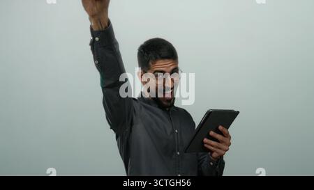 Young man with glasses gestures energetically while holding a tablet against a white background. Stock Photo
