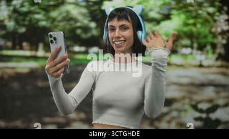 Young hispanic woman with cat ear headphones holds smartphone and beckons hand in forest; friendly invitation. Stock Photo