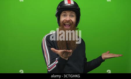 Young man with long beard wearing black motorcycle helmet and striped sleeve sweater presents open palms in green studio; joy. Stock Photo