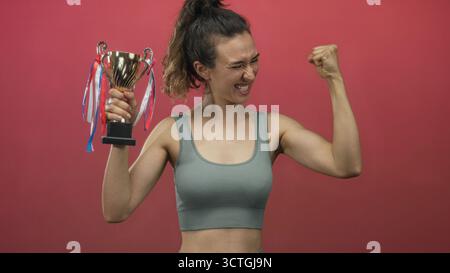Woman holds trophy with fist raised in studio against a vivid red backdrop, smiling broadly; pride celebration. Stock Photo