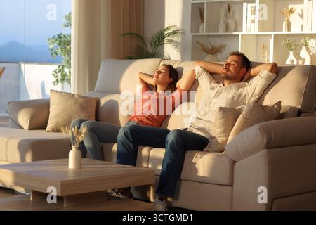 Happy couple relaxing sitting on a couch with arms on head at sunset at home Stock Photo