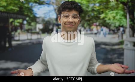 Man wearing round glasses and a white sweater shrugs both shoulders amid passersby on a sunny urban street; uncertainty. Stock Photo