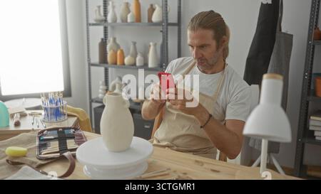 Man with long blond hair and apron holds red smartphone while seated at pottery wheel and examines a clay vase in studio; creative focus. Stock Photo