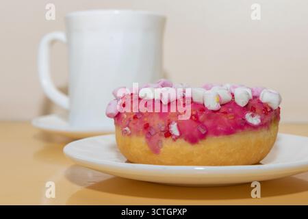 Donut with pink icing and marshmallows on plate with white cup on table. Sweet dessert for coffee break. Stock Photo