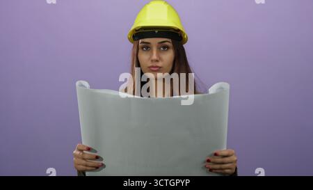 Young woman architect wearing yellow hardhat examines blueprint against purple background with focus and concentration, showcasing engineering skills Stock Photo