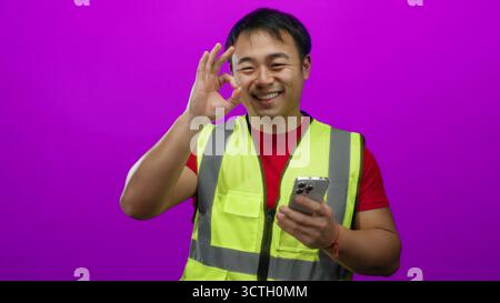 Man wearing safety vest using smartphone against vibrant pink background looks focused while texting an important message, embodying modern communicat Stock Photo