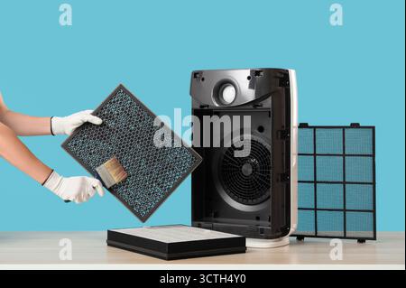 Female technician with brush cleaning dirty air purifier filter on blue background Stock Photo