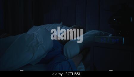Man in Blue Pajamas Goes Bed, Digital Alarm Clock Shows 3 AM. Tired From Insomnia, Man Tries Fall Asleep in Early Hours of Morning. Concept Sleep Deprivation, Silent Struggle With Nighttime Anxiety. Stock Photo