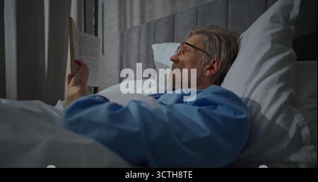 Waking Up in Morning in Bright, Cozy Bedroom, Mature Man Wearing Glasses Lies Relaxed in White Bed Reading Book. Pleasant, Leisurely Morning, Relaxing in Bed. Concept of Morning Routine. Static Shot. Stock Photo
