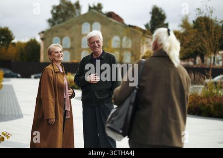 Senior Caucasian man and elderly woman standing outdoors smiling and talking with female friend approaching outdoors, facing away, engaging in friendly conversation while walking in urban park Stock Photo