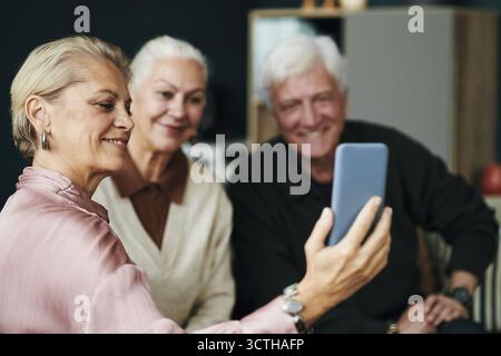 Senior Caucasian woman taking selfie with smartphone while sitting with male and female friend couple, all smiling and looking at device together while taking photo for social media Stock Photo