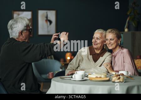 Senior Caucasian man photographing senior woman and middle aged female friend sitting together at table smiling, enjoying dessert and drinks in cozy indoor setting Stock Photo