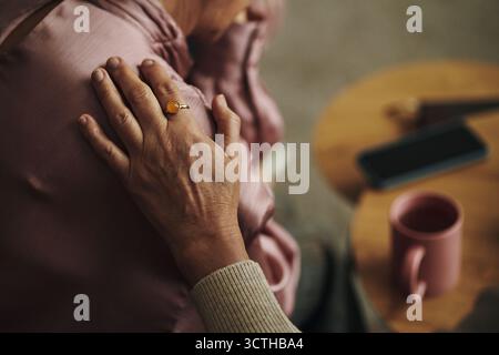 Elderly woman comforting senior female friend by gently placing hand on shoulder, woman sitting with head resting on hand near smartphone and coffee mug in living room Stock Photo