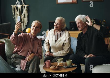 Three Caucasian seniors sitting together, smiling and posing for selfie with smartphone, older man waving hand while two senior women looking at camera, drinks and snacks on table Stock Photo