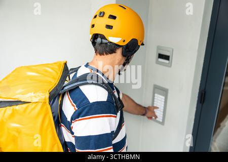 Hispanic delivery courier in yellow helmet and insulated backpack pressing an outdoor intercom button at an apartment entrance, providing fast, contac Stock Photo