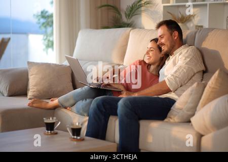 Happy couple at home using laptop sitting on a couch at sunset Stock Photo