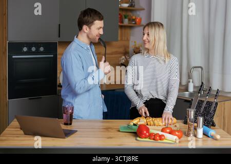 Home concert. Playful spouses having fun in kitchen, singing and dancing. Cheerful couple using kitchen utensils as microphones, fooling while cooking Stock Photo