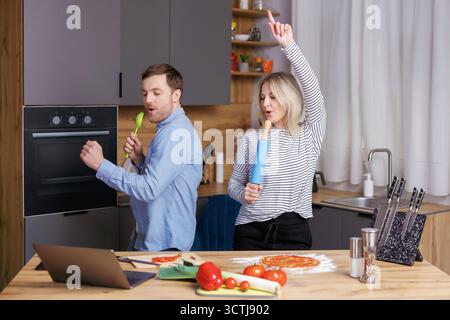 Home concert. Playful spouses having fun in kitchen, singing and dancing. Cheerful couple using kitchen utensils as microphones, fooling while cooking Stock Photo