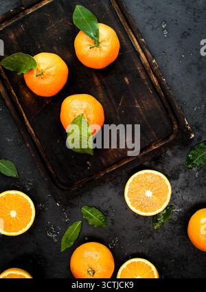 Whole and halved fresh oranges with leaves arranged vertically on a dark surface, captured in bright side light for vibrant citrus imagery. Stock Photo