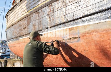 Man wearing a green cap and long sleeve shirt with gloves, tarring wads of hemp in a gap on the hull of a wooden boat Stock Photo