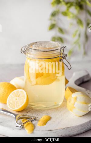 Glass jar of lemon peels steeping in alcohol with peeled lemons and a zester on a white board, bright and fresh. Stock Photo