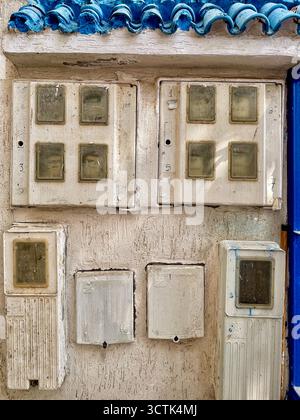Weathered electrical meter boxes mounted on textured wall beneath blue tiled roof edge, Martil, Tetouan, Morocco, 6 October 2025. Stock Photo