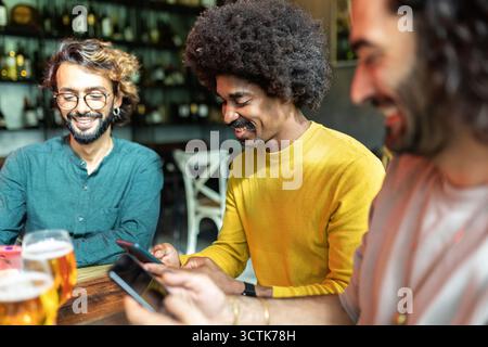 Three diverse young adult friends using mobile phone sitting on table at bar Stock Photo