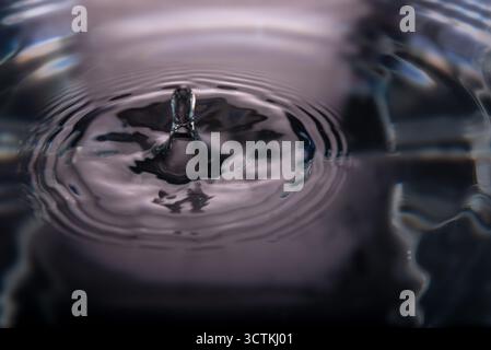 186.Water droplet rebounds above surface forming vertical column. Concentric ripples and reflections on glossy water highlight fluid motion and surfac Stock Photo