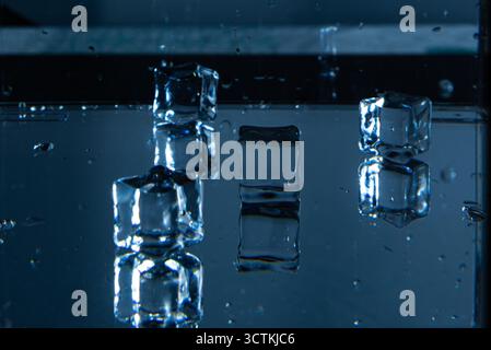 4.Five clear ice cubes rest on a reflective surface with scattered water droplets. The bluish lighting and blurred background emphasize texture, symme Stock Photo