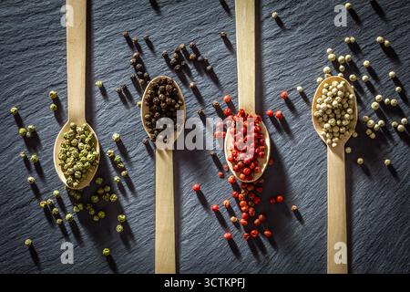 Four wooden spoons filled with green, black, red, and white peppercorns on slate—an elegant study in spice, texture, and culinary color. Stock Photo