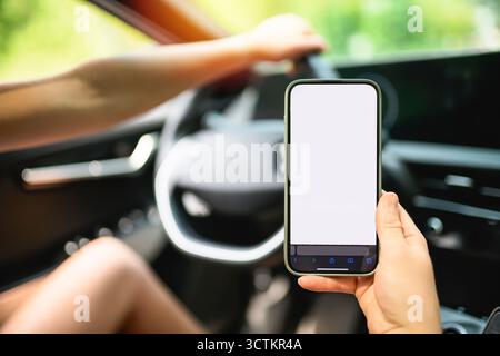 Close-up of female hand holding smartphone with white mockup on screen, background of car steering wheel. Stock Photo