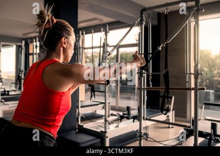 Woman seen from behind exercising and toning her arms on a Pilates reformer machine during a beautiful golden hour sunset in a modern gym. Stock Photo