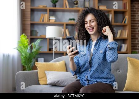 Curly-haired woman sitting on a couch at home, cheering and feeling excited, successfully looking at her smartphone and celebrating a victory or achieving a goal Stock Photo