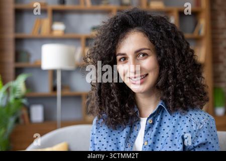 Smiling young woman with natural curly hair and brown eyes poses confidently in a cozy modern living room, radiating authenticity, warmth and relaxed professional charm Stock Photo