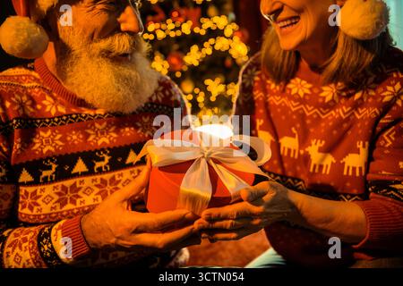 Couple in festive sweaters exchange a gift box with a white ribbon during Christmas night at home creating warm holiday joy and family feeling Stock Photo