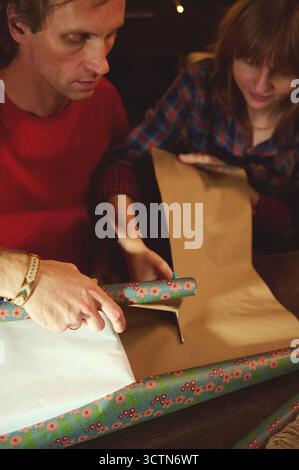 Two adults wrap gifts with floral paper in a warm, intimate home scene. Focus on hands, tape, and the care of preparing presents for family and friend Stock Photo