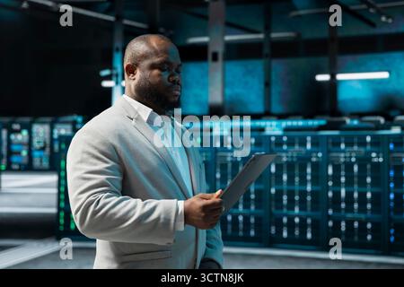 Programmer in data center updating systems using tablet, debugging app performance issues. Server hub IT expert using device, looking for malfunctions, increasing maximum efficiency and uptime Stock Photo