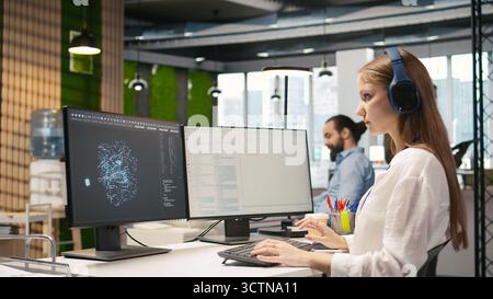 Female technician coding with AI language model assistance in startup office, listening music. Young woman wearing headphones integrating LLM assistant suggestions into code editor, camera A Stock Photo