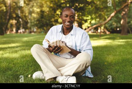 Happy african american guy studying with book and laptop in park, sitting in college campus and taking notes Stock Photo