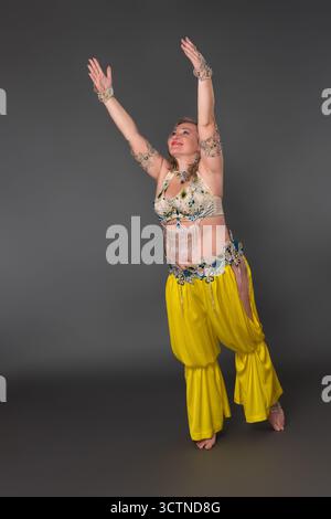 Happy and smiling woman dancer with raised arms wearing in dancing costume - top and yellow harem trousers, moves barefoot in full length against gray Stock Photo