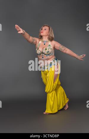 Caucasian woman dancer with outstretched arms performing in full length against gray background. Woman dressing in dancing costume - top and yellow ha Stock Photo