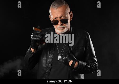 Stylish old man in leather jacket with cigar on black background Stock Photo