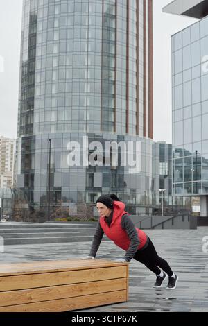 Young sporty Caucasian woman doing push-ups on wooden bench while having outdoor training in city center on cold autumn day Stock Photo